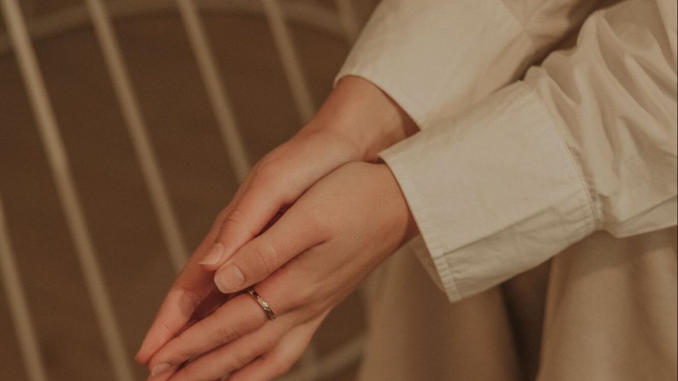Plano detalhado das mãos de uma pessoa sentada, com os dedos entrelaçados suavemente sobre as pernas, vestindo roupa clara em tons bege e branco.